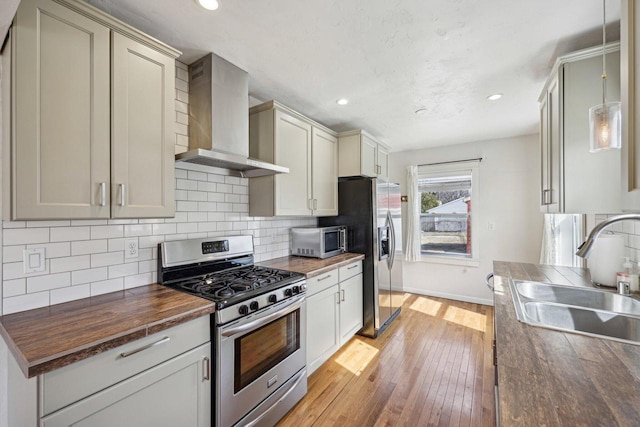 kitchen with wooden counters, wall chimney range hood, light wood-type flooring, appliances with stainless steel finishes, and a sink