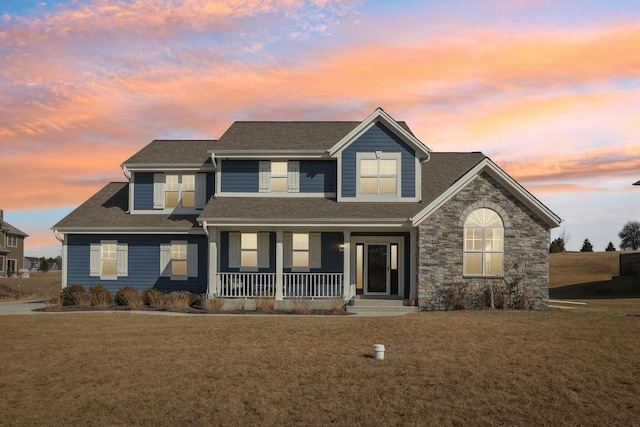 view of front of property featuring a yard, stone siding, covered porch, and a shingled roof