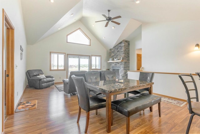 dining room featuring baseboards, light wood finished floors, high vaulted ceiling, ceiling fan, and a stone fireplace