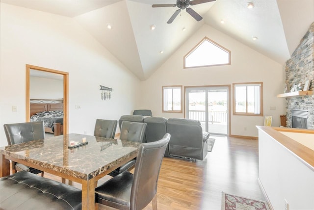 dining space with a stone fireplace, high vaulted ceiling, light wood-type flooring, and ceiling fan