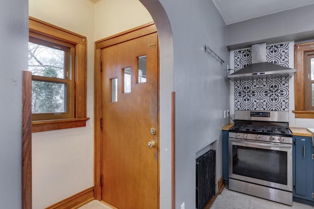 kitchen with baseboards, blue cabinetry, arched walkways, gas range, and wall chimney exhaust hood