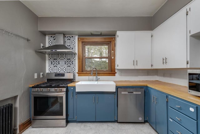 kitchen featuring blue cabinetry, a sink, stainless steel appliances, radiator, and wall chimney exhaust hood