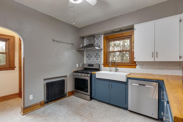 kitchen featuring blue cabinetry, a sink, stainless steel appliances, radiator, and wall chimney range hood