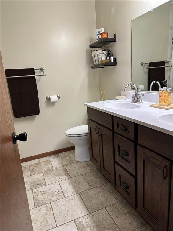 bathroom featuring toilet, a sink, stone tile flooring, double vanity, and baseboards