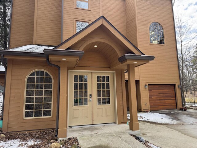 snow covered property entrance with an attached garage, french doors, and concrete driveway