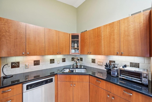 kitchen featuring tasteful backsplash, dark stone countertops, stainless steel dishwasher, a sink, and glass insert cabinets