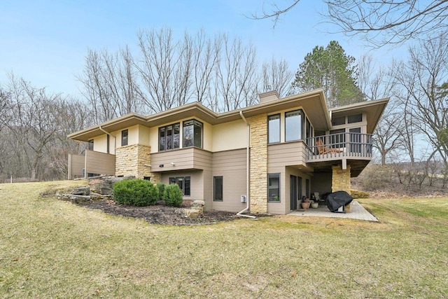 rear view of property featuring stone siding, a balcony, a patio, a chimney, and a lawn
