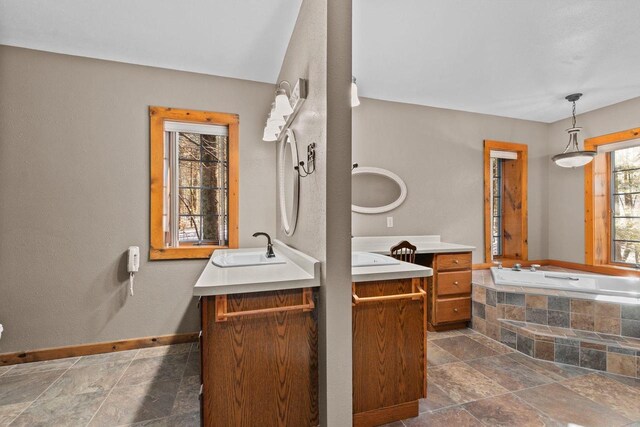 bathroom with tiled tub, stone finish flooring, baseboards, two vanities, and a sink