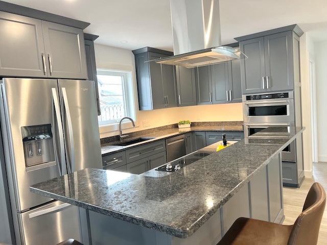 kitchen with island exhaust hood, dark stone countertops, stainless steel appliances, light wood finished floors, and a sink