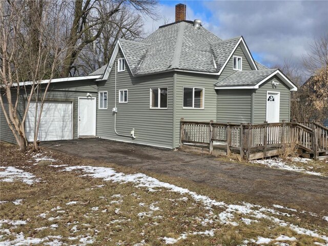 snow covered back of property with a shingled roof, driveway, a garage, and a chimney