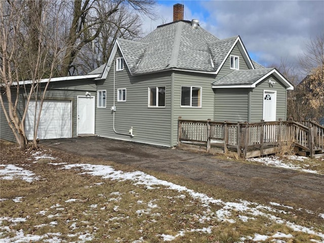 snow covered back of property with an attached garage, a shingled roof, driveway, and a chimney