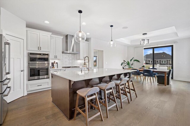 kitchen with wall chimney range hood, dark wood-style floors, a tray ceiling, stainless steel appliances, and a sink
