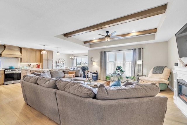 living room with a glass covered fireplace, light wood-style floors, ceiling fan with notable chandelier, and beamed ceiling