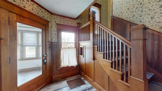 entryway featuring light wood-type flooring, ornamental molding, a wainscoted wall, stairs, and wallpapered walls