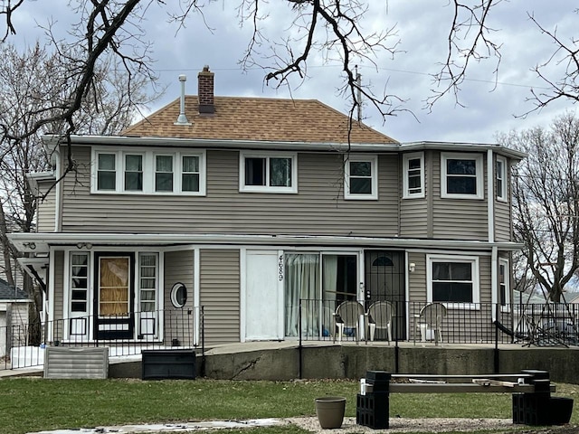 rear view of property featuring roof with shingles, a porch, a lawn, and a chimney