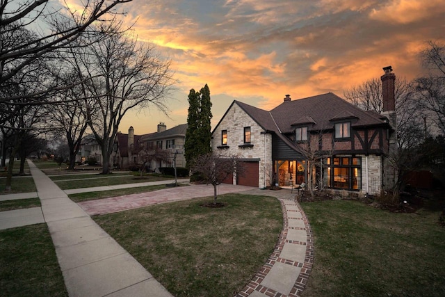 french country style house with a garage, stone siding, a chimney, a lawn, and driveway