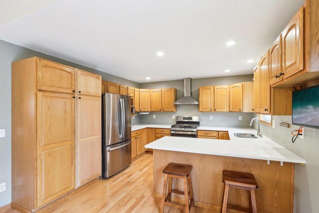 kitchen with a peninsula, light wood-style floors, wall chimney range hood, a sink, and appliances with stainless steel finishes