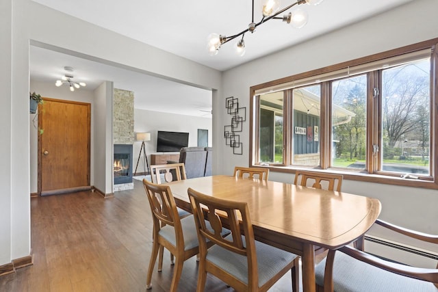 dining space with a stone fireplace, wood finished floors, baseboards, a baseboard heating unit, and a notable chandelier