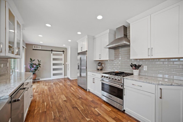 kitchen featuring a barn door, backsplash, wall chimney exhaust hood, appliances with stainless steel finishes, and visible vents