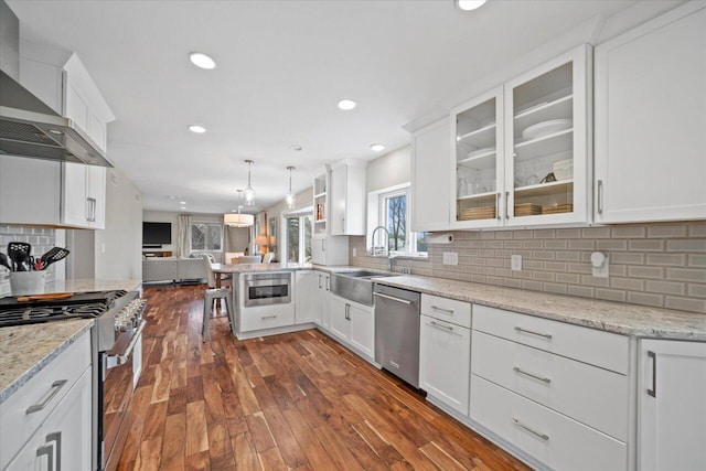 kitchen with white cabinets, stainless steel appliances, wall chimney range hood, a sink, and open floor plan