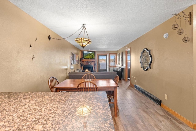 dining room with a lit fireplace, a textured ceiling, wood finished floors, a baseboard heating unit, and baseboards