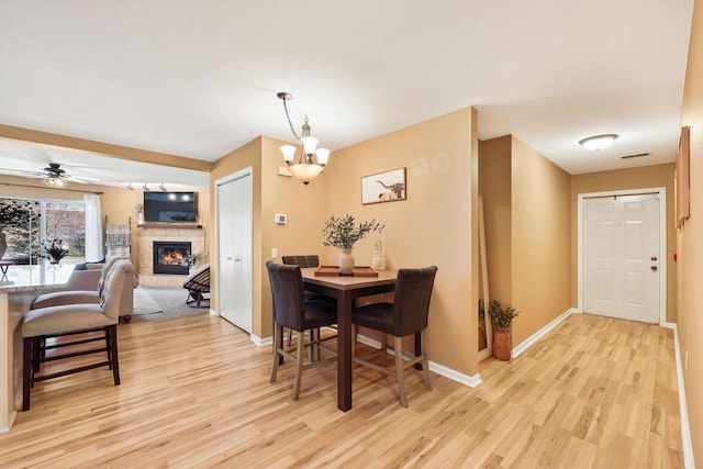 dining space with visible vents, light wood-style floors, baseboards, a tiled fireplace, and ceiling fan with notable chandelier