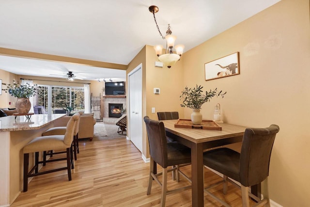 dining space featuring baseboards, light wood-style flooring, ceiling fan with notable chandelier, and a fireplace