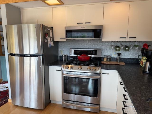 kitchen with light wood finished floors, dark stone countertops, tasteful backsplash, white cabinetry, and appliances with stainless steel finishes