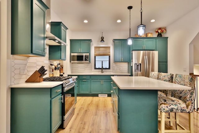 kitchen with wall chimney range hood, light countertops, light wood-style flooring, a kitchen bar, and appliances with stainless steel finishes