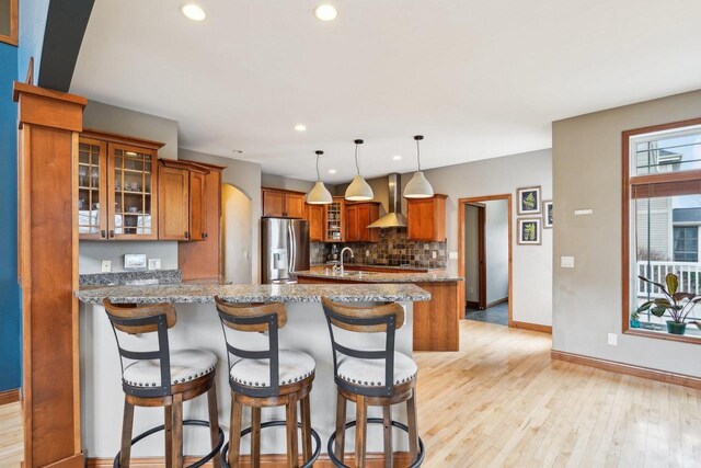 1310 Hanover Ct, Waunakee, WI 53597 - kitchen with wall chimney exhaust hood, a peninsula, baseboards, stainless steel fridge with ice dispenser, and brown cabinetry