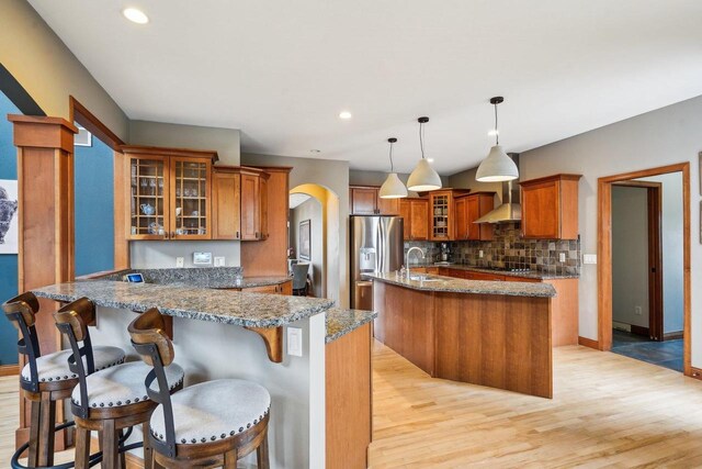 1310 Hanover Ct, Waunakee, WI 53597 - kitchen with wall chimney range hood, light stone counters, brown cabinetry, stainless steel fridge, and black electric cooktop