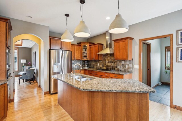 1310 Hanover Ct, Waunakee, WI 53597 - kitchen with arched walkways, a sink, brown cabinets, wall chimney range hood, and appliances with stainless steel finishes