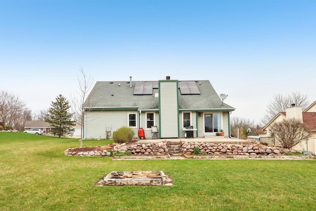 rear view of house with a patio area, a lawn, a chimney, solar panels, and roof with shingles