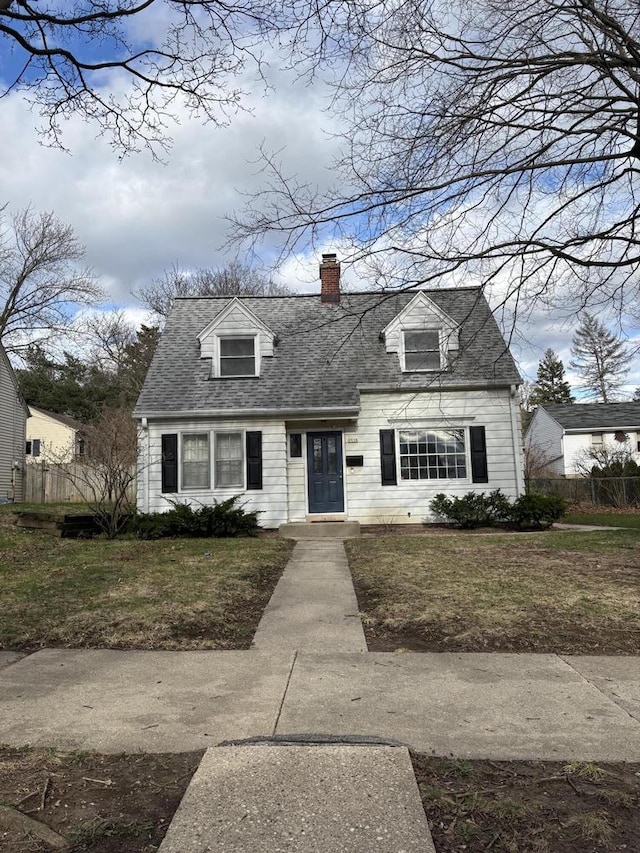 cape cod house with a chimney, roof with shingles, fence, and a front yard