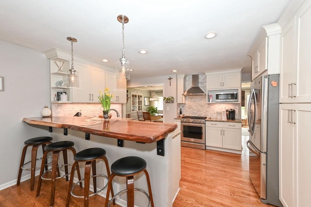 kitchen with wall chimney range hood, wood counters, stainless steel appliances, a kitchen bar, and backsplash