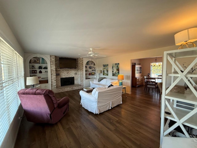 living area featuring a fireplace, built in features, dark wood-style floors, and ceiling fan with notable chandelier