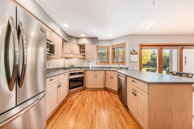 kitchen featuring light wood finished floors, stainless steel appliances, a peninsula, a sink, and light brown cabinets