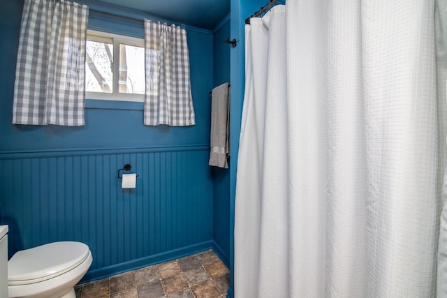 full bathroom with wainscoting, stone finish floor, toilet, and a shower with curtain