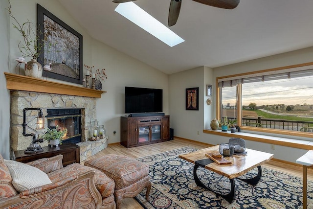 living area featuring vaulted ceiling with skylight, ceiling fan, a stone fireplace, wood finished floors, and baseboards