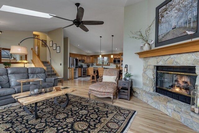 living room featuring a ceiling fan, a stone fireplace, high vaulted ceiling, light wood-style floors, and stairs