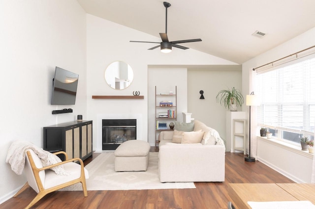 living area with ceiling fan, a glass covered fireplace, visible vents, vaulted ceiling, and dark wood finished floors
