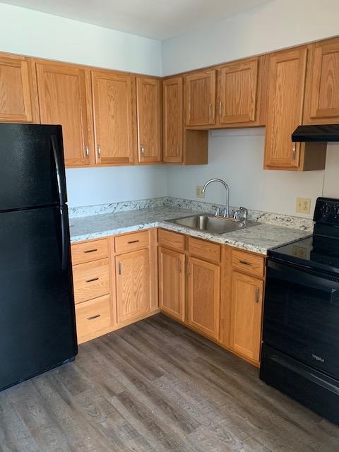 kitchen with light stone counters, a sink, under cabinet range hood, black appliances, and dark wood finished floors