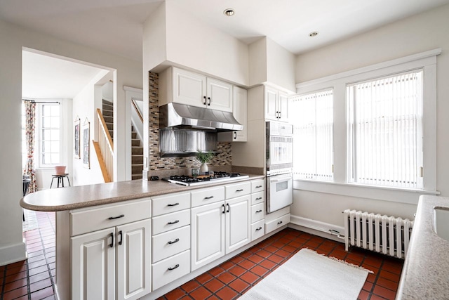 kitchen featuring decorative backsplash, under cabinet range hood, radiator heating unit, dark tile patterned flooring, and white appliances