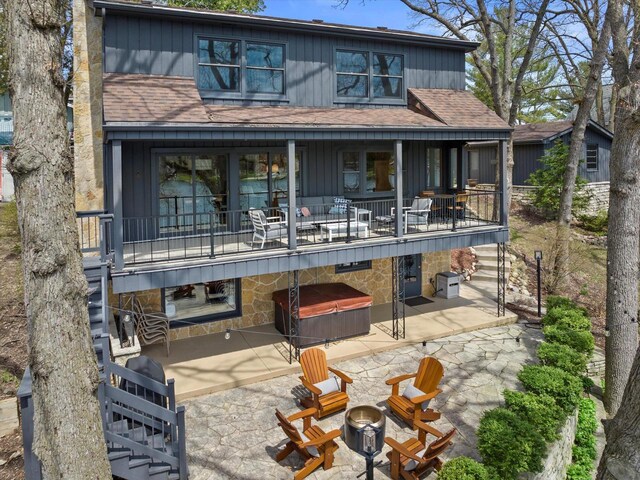 back of property with a balcony, stone siding, a patio area, a hot tub, and roof with shingles