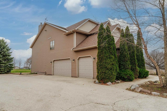 view of side of home with driveway, a chimney, an attached garage, and roof with shingles