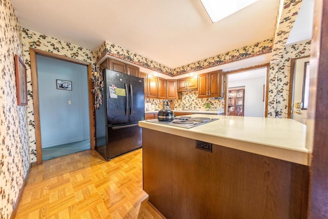 kitchen with freestanding refrigerator, wallpapered walls, light countertops, brown cabinets, and stainless steel stovetop