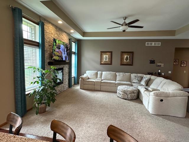 carpeted living room with visible vents, a raised ceiling, ceiling fan, and a stone fireplace