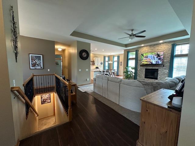 living area featuring a ceiling fan, a stone fireplace, wood finished floors, and a raised ceiling