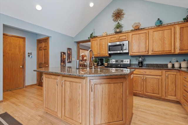 kitchen featuring dark stone counters, a sink, stainless steel appliances, light wood-style floors, and a center island with sink