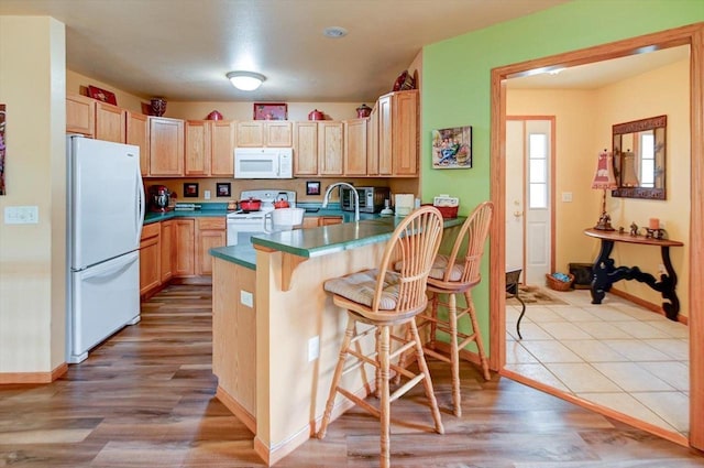 kitchen featuring white appliances, a peninsula, light brown cabinetry, and wood finished floors